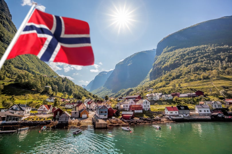 Natural Pharmaceuticals Fleximed® Active Norra küla fjordi ääres, ümbritsetud mägedest ja rohelusest, Norra lipu lehvimas esimesel plaanil selge, päikesepaistelise taeva all.