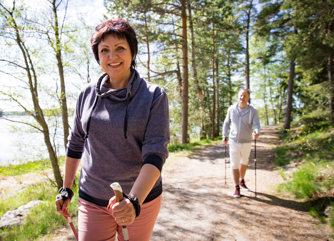 Natural Pharmaceuticals FlexiMed® Active Naeratav naine ja mees matkavad kepikõndi tehes päikeselisel metsateel rohelises looduses.