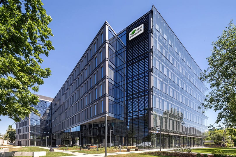 Natural Pharmaceuticals FlexiMed® Active Kaasaegne klaasfassaadiga kontorihoone roheliste puudega pargi taustal, selge sinise taeva all.